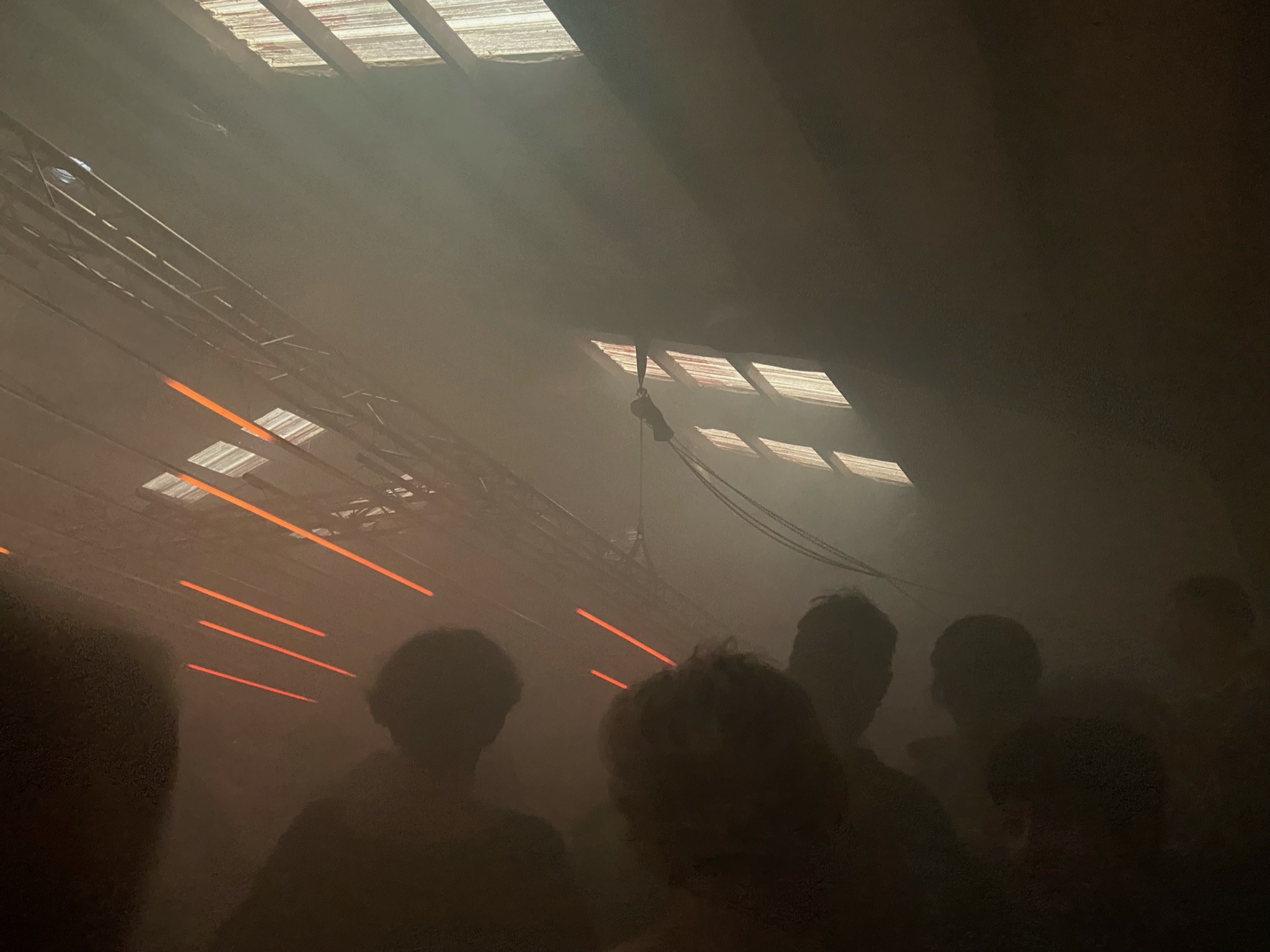 A crowded live venue beneath an industrial ceiling, lit by warm haze and red bars.